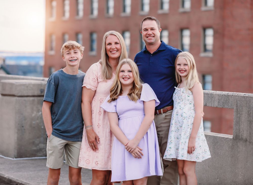 A family of five poses together on a rooftop terrace with a brick building backdrop at sunset. Best family photographer Peoria, Il.