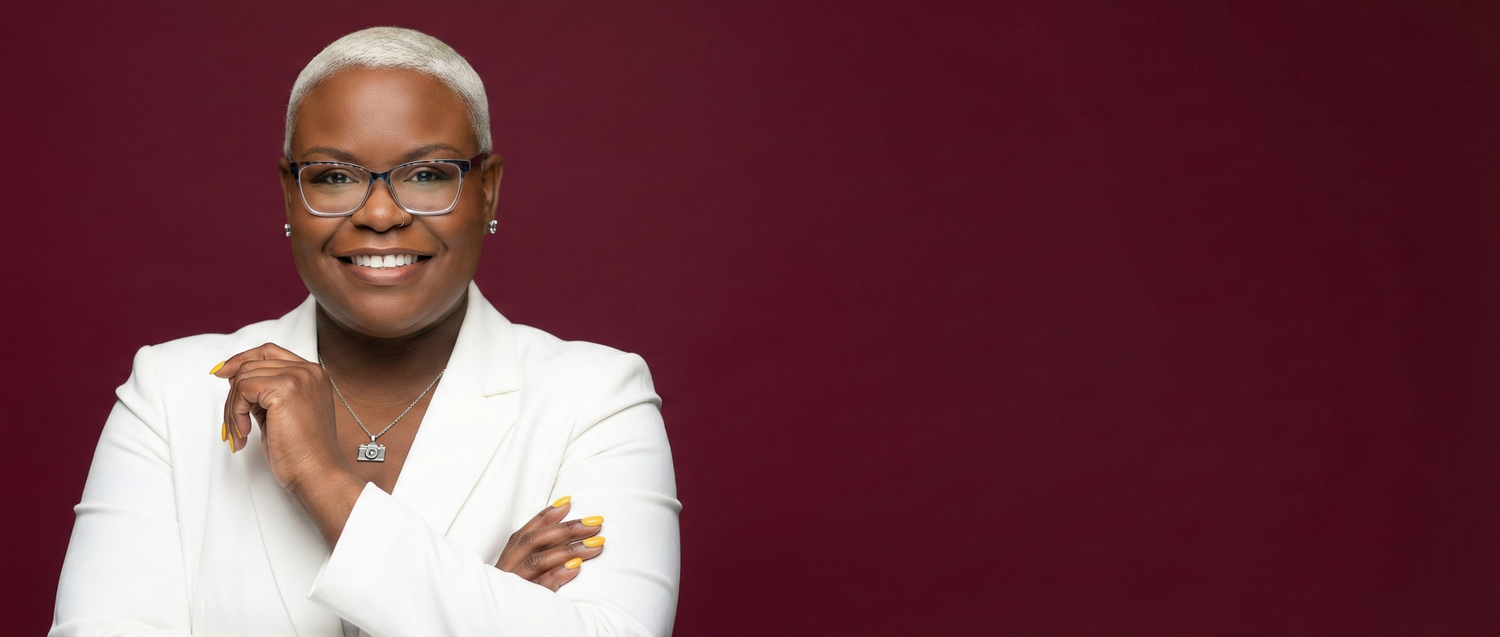 A person in a white suit jacket poses confidently against a dark burgundy background.