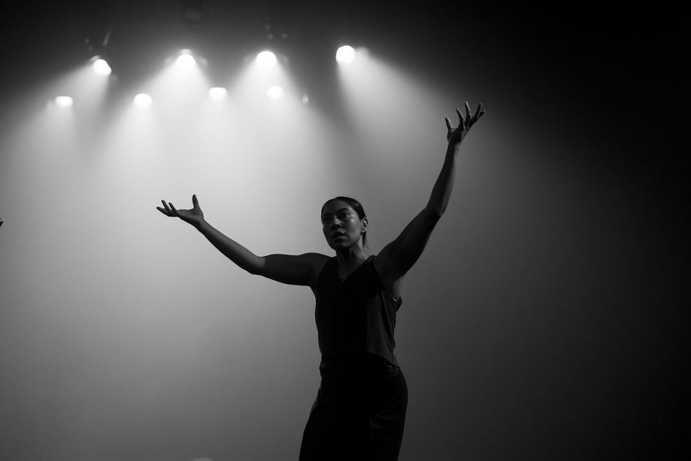 Dramatic black and white performance with figures reaching upward against bright stage lights.