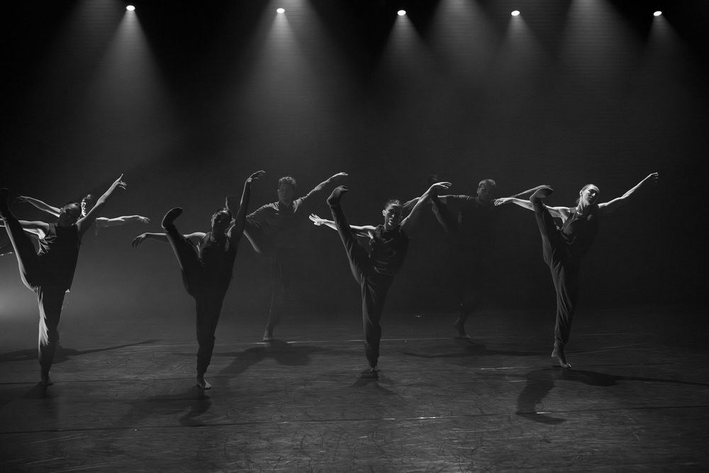Dancers perform a synchronized movement sequence on a dimly lit stage with dramatic spotlighting.