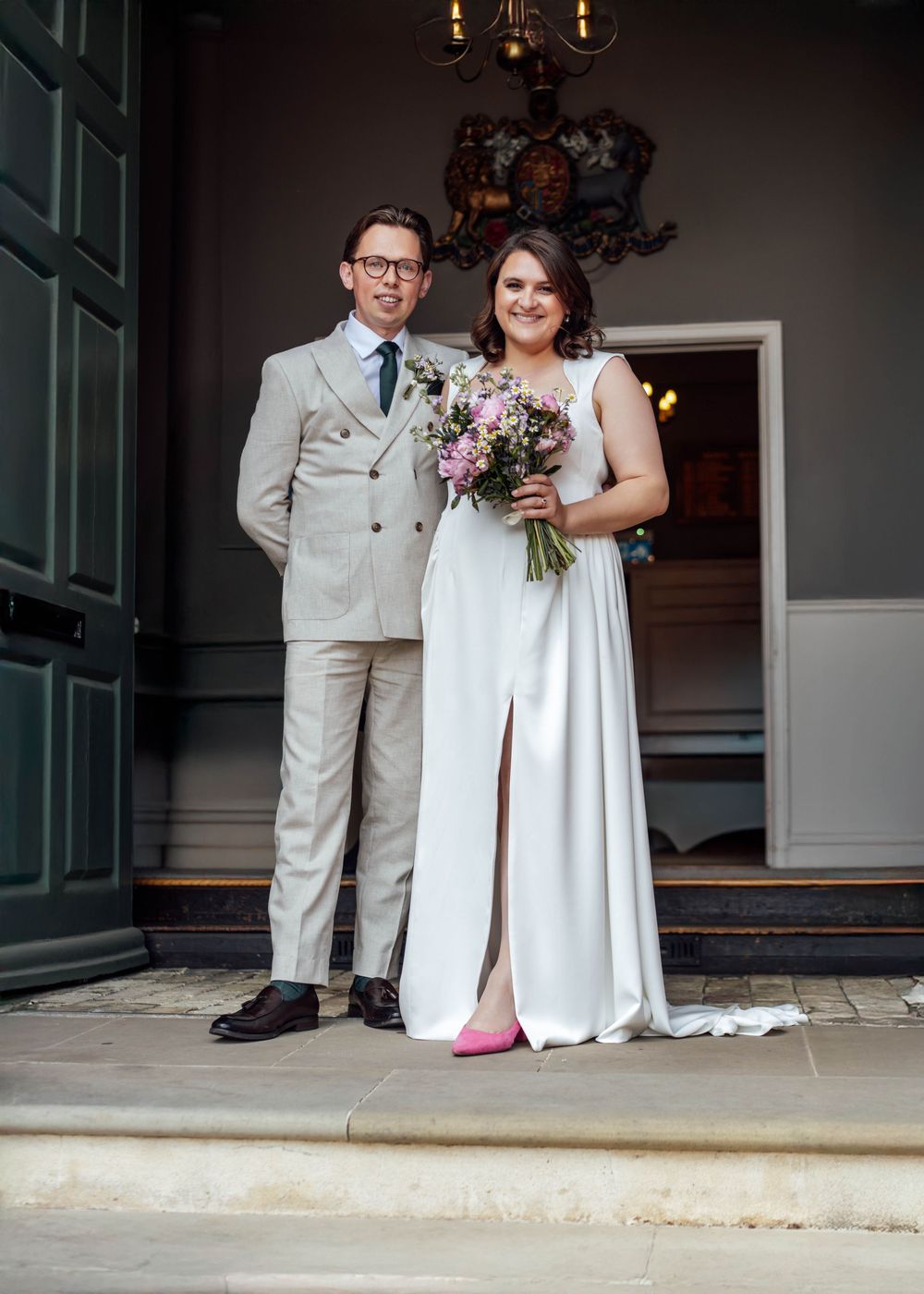 Bride in white gown with pink shoes holding floral bouquet stands with groom in beige suit outside elegant venue doors. London Wedding