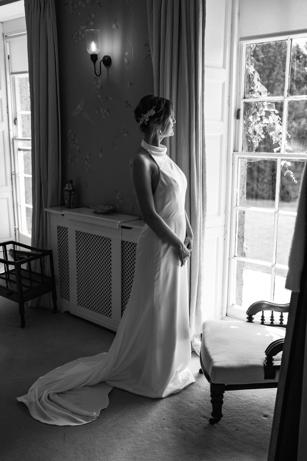 Bride in elegant white halter gown with train stands by window in black and white wedding portrait. London Wedding Photographer