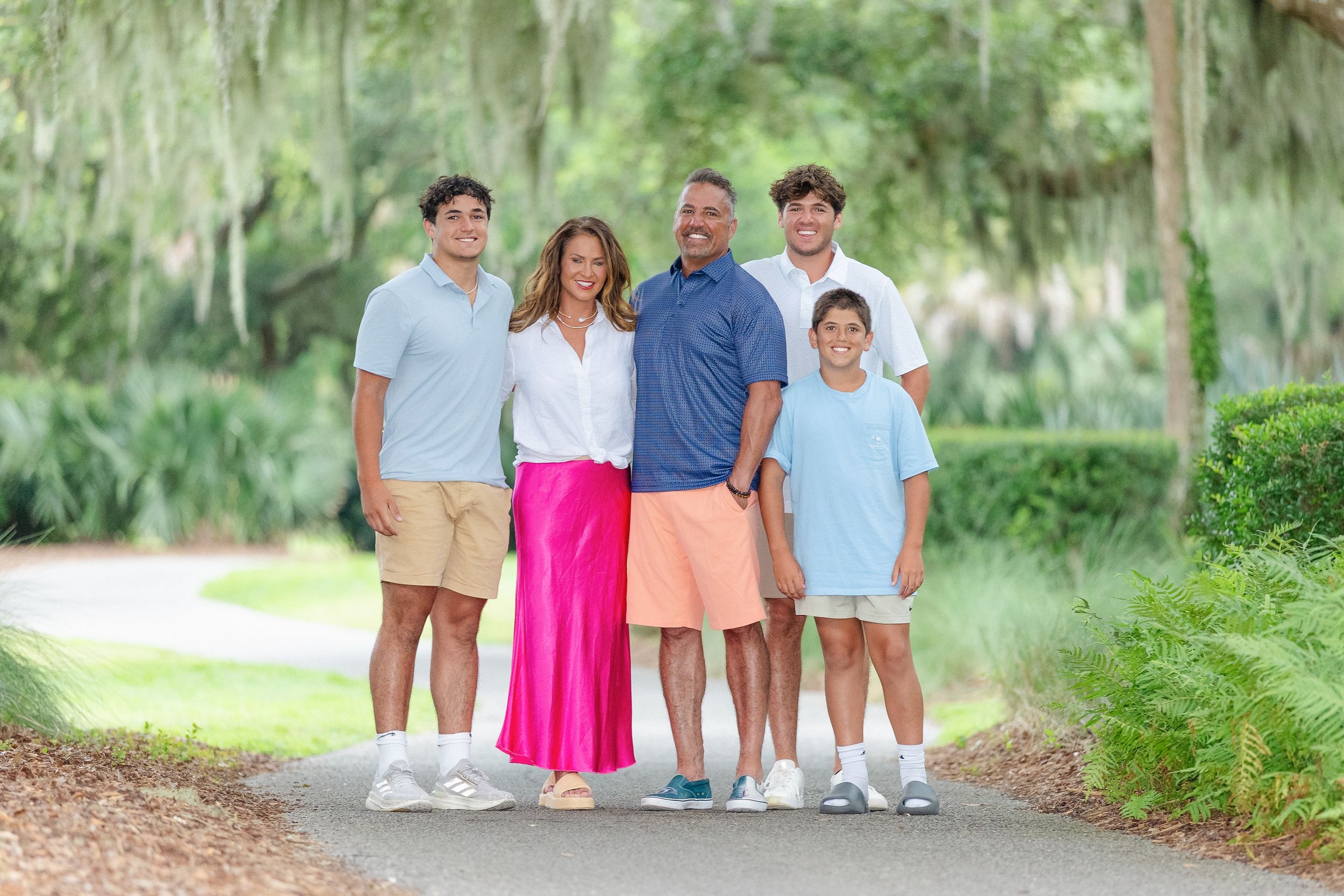Family of five posing together on a sunlit path surrounded by moss-covered trees in a park.