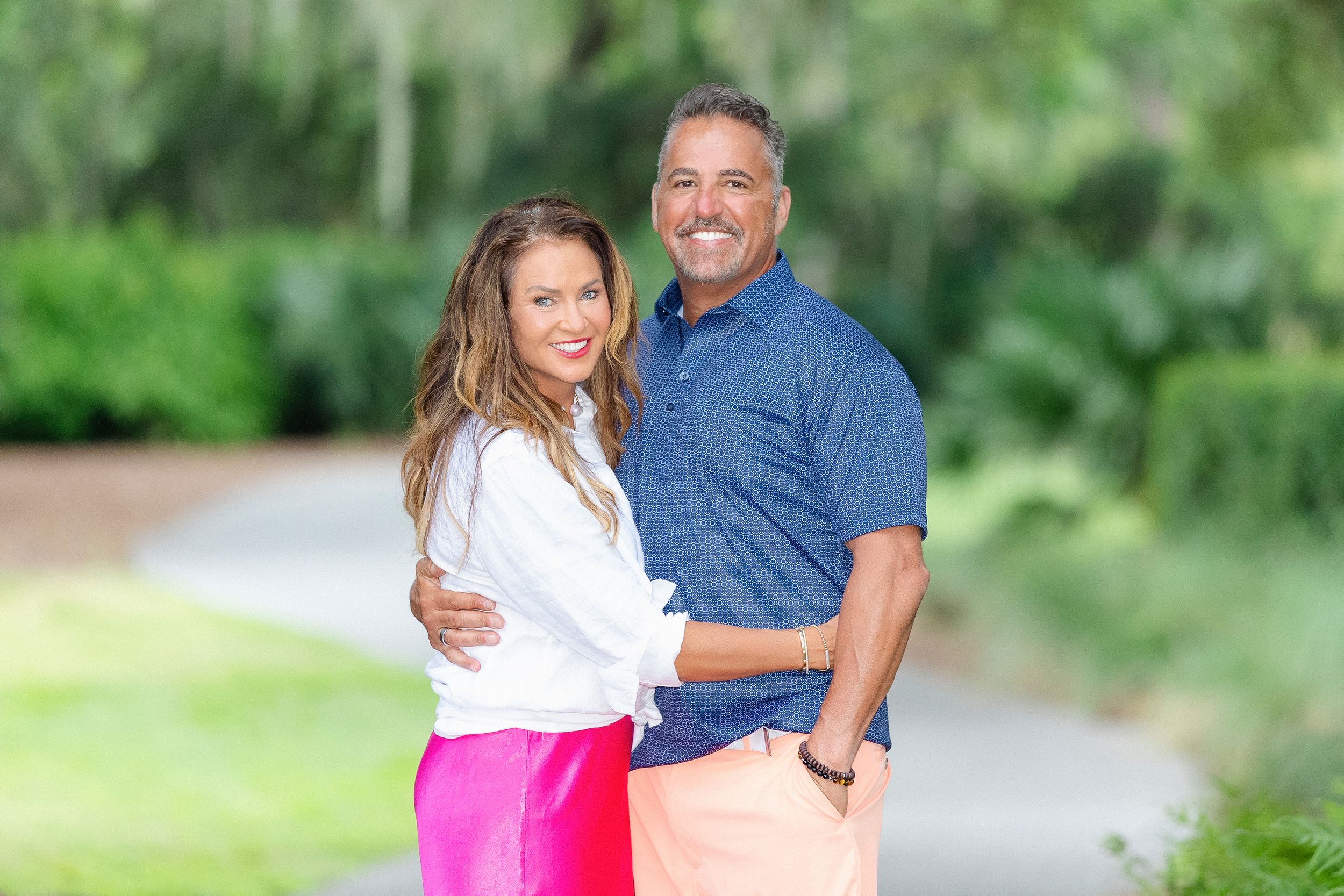 Smiling couple posing together on a lush green tree-lined path on a sunny day.