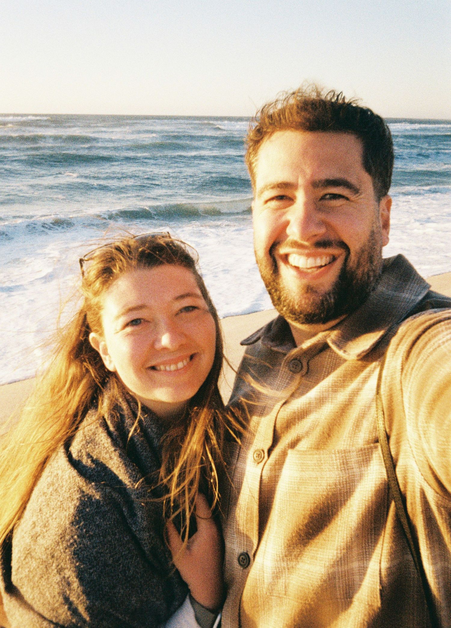 A couple takes cheerful selfies together at the beach during golden hour with the ocean visible in the background.