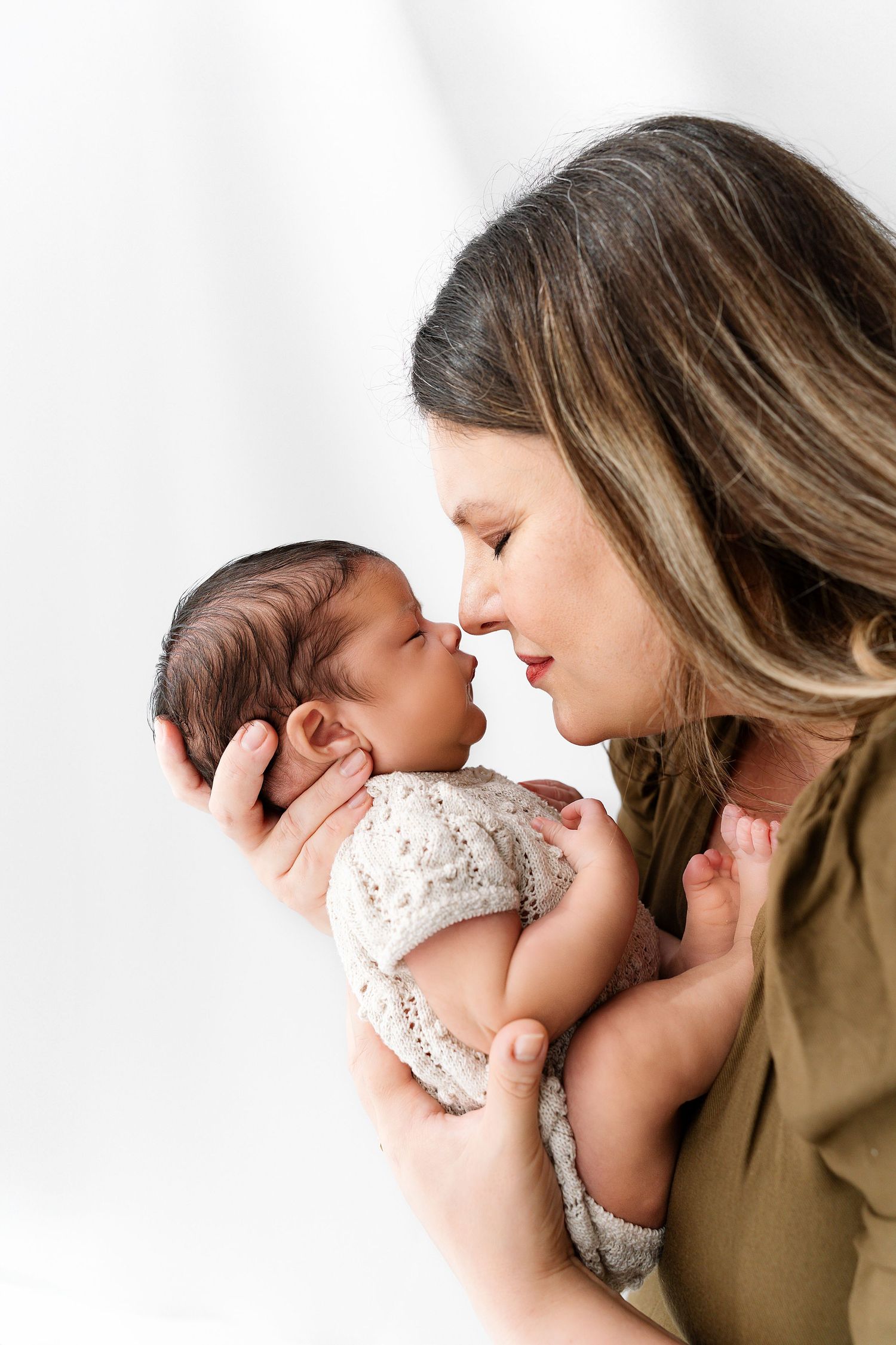 A tender moment between mother and newborn baby as they touch noses against a white background.
