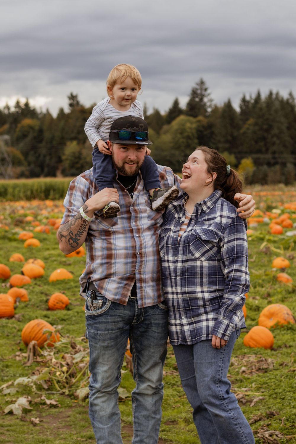 Smiling family at pumpkin patch, dad carrying toddler on shoulders while mom laughs beside them.