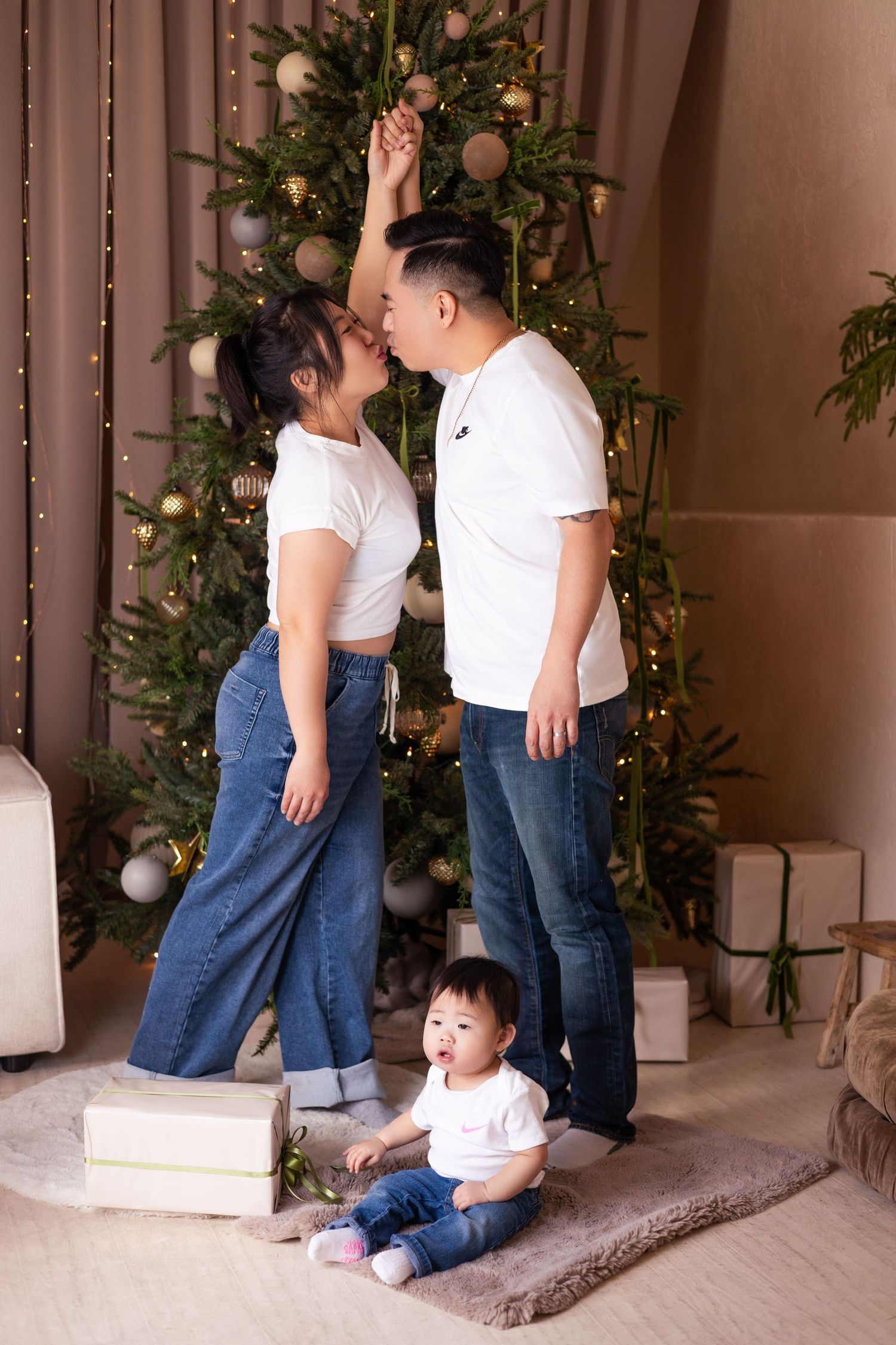 Asian family in white tees and jeans sharing a kiss under mistletoe beside a decorated Christmas tree.