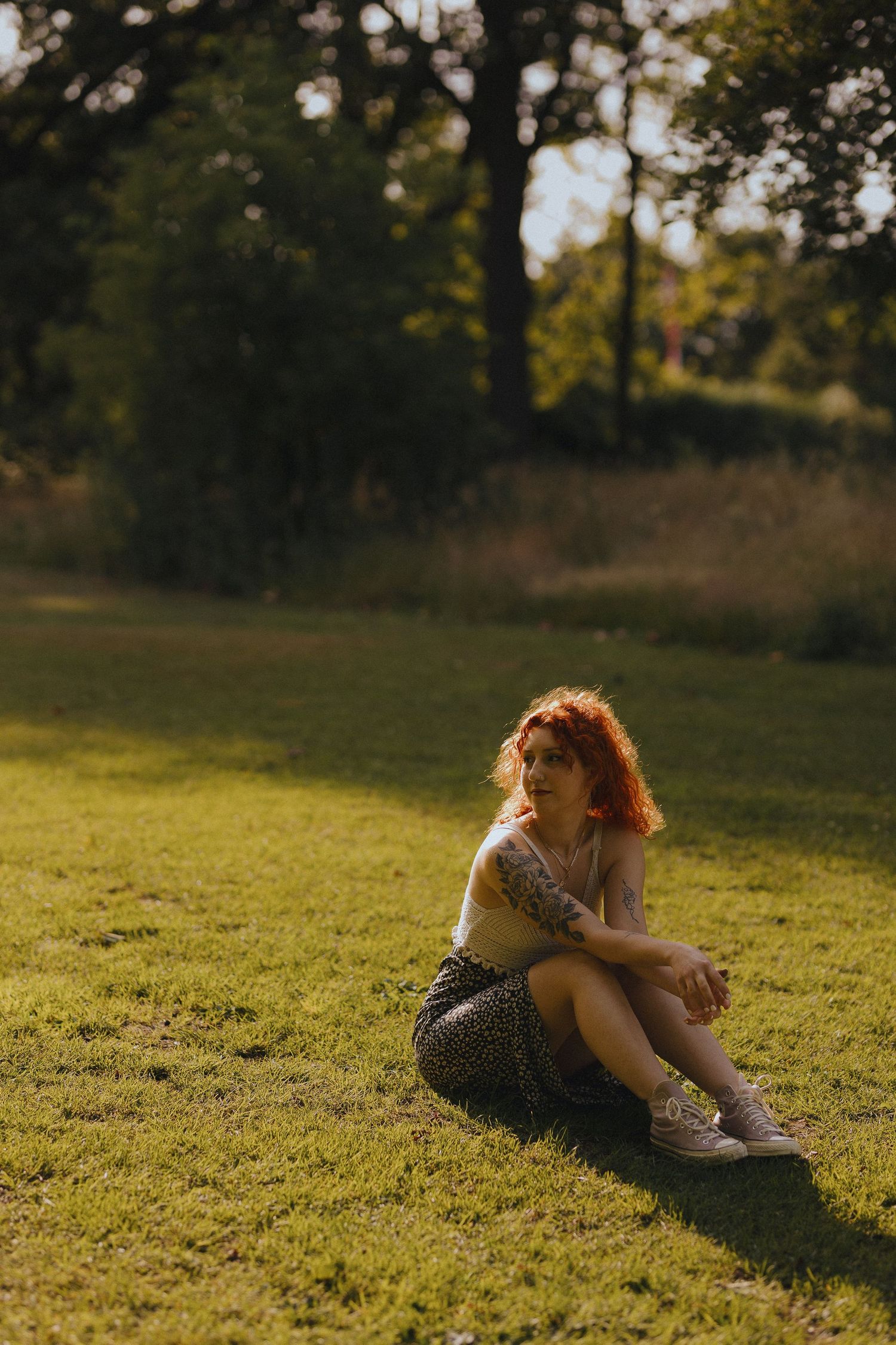 A person with vibrant red curly hair twirls playfully in a polka dot dress and olive jacket outdoors in a park setting.