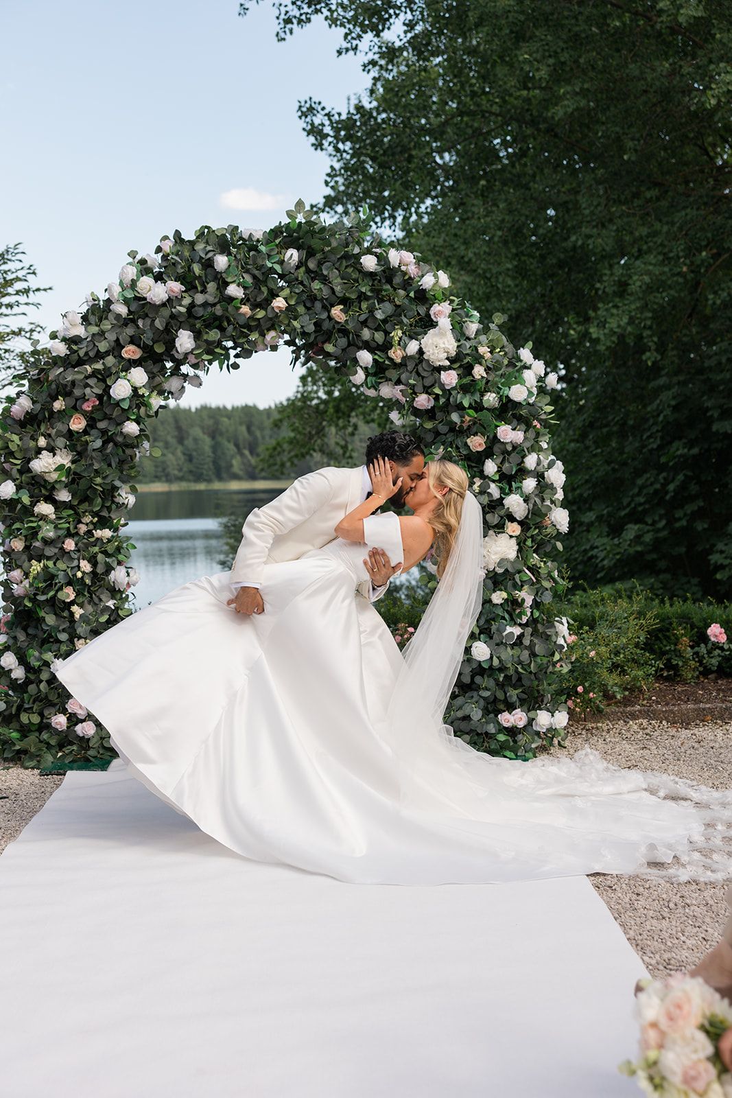 A romantic wedding portrait by a lake with a floral arch and flowing white dress billowing in the breeze.