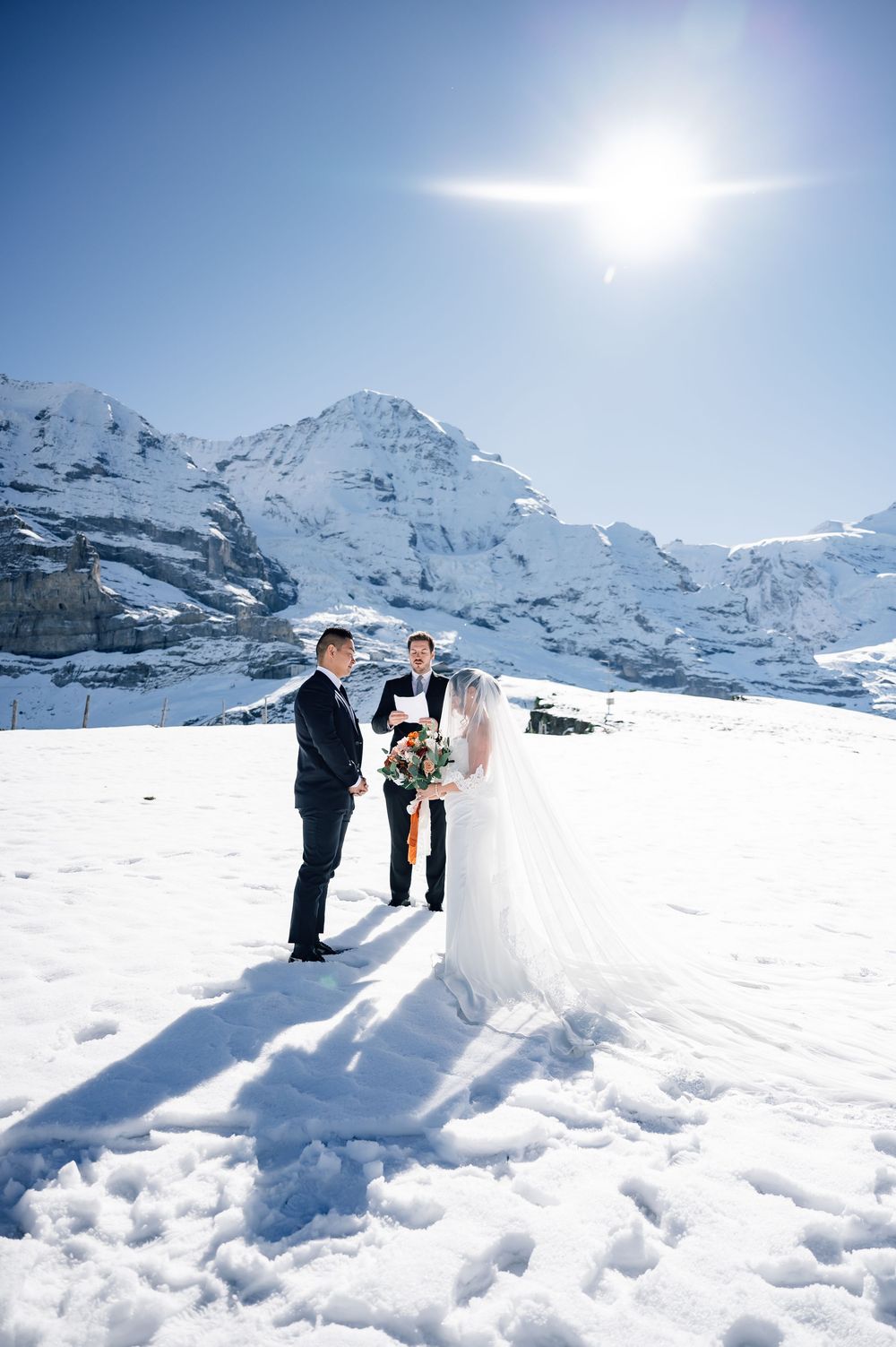 A winter wedding ceremony takes place on a snow-covered mountain peak with majestic snow-capped Alps in the background.