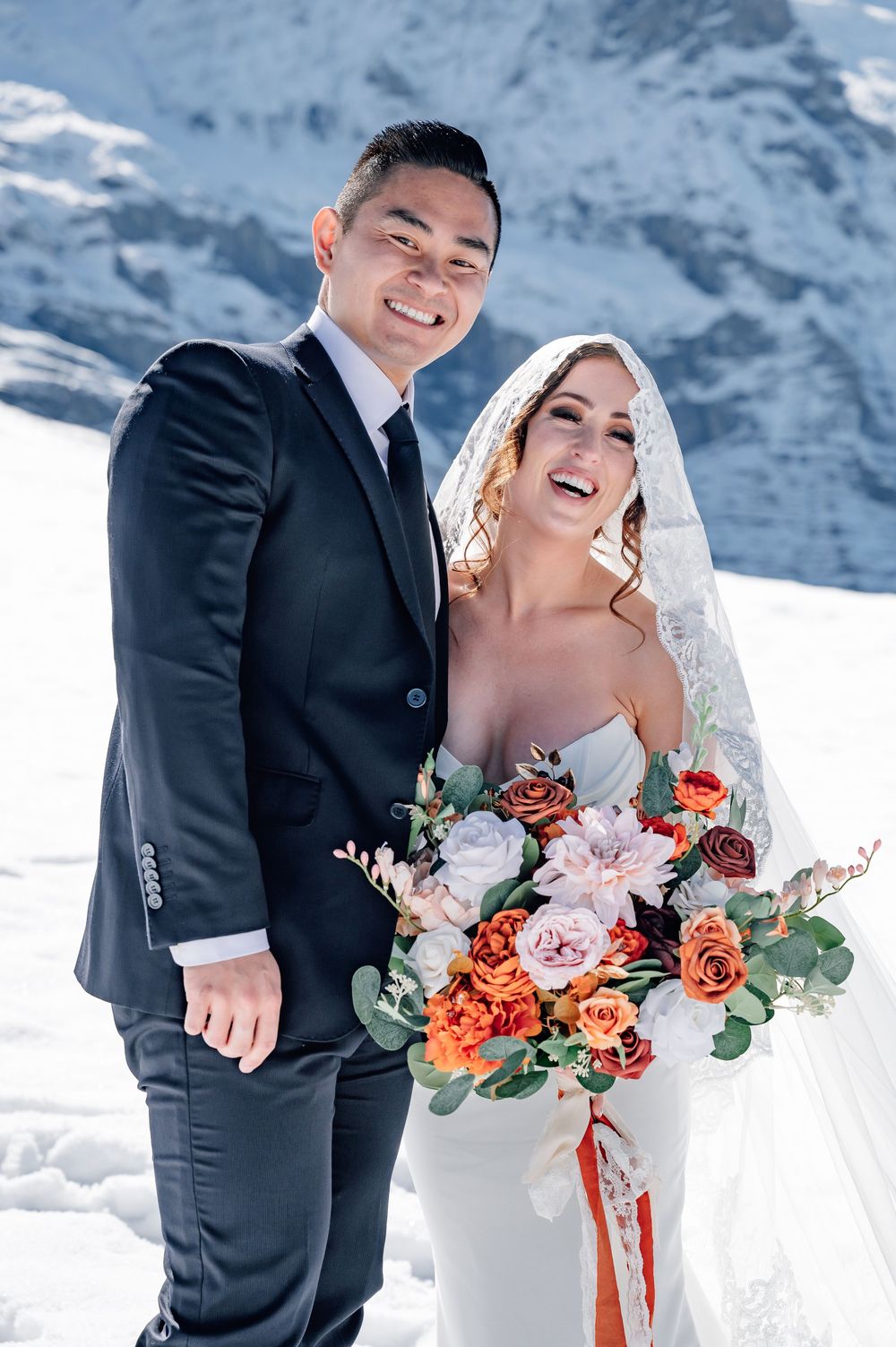 Wedding couple shares joyful moments against snowy mountain backdrop with vibrant coral and pink floral bouquet.