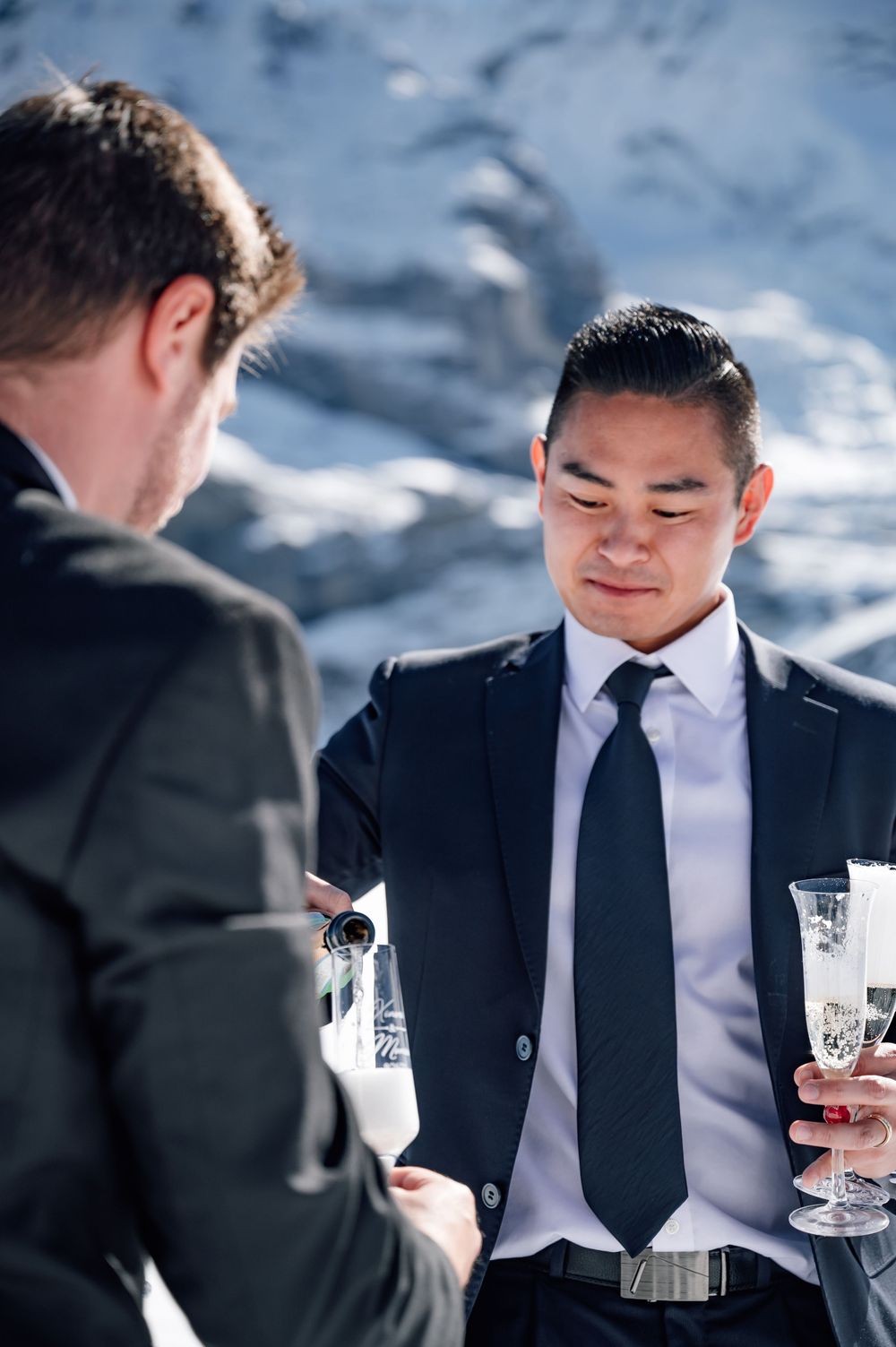 Two people in formal attire sharing drinks at an outdoor event with a blurred mountain background.