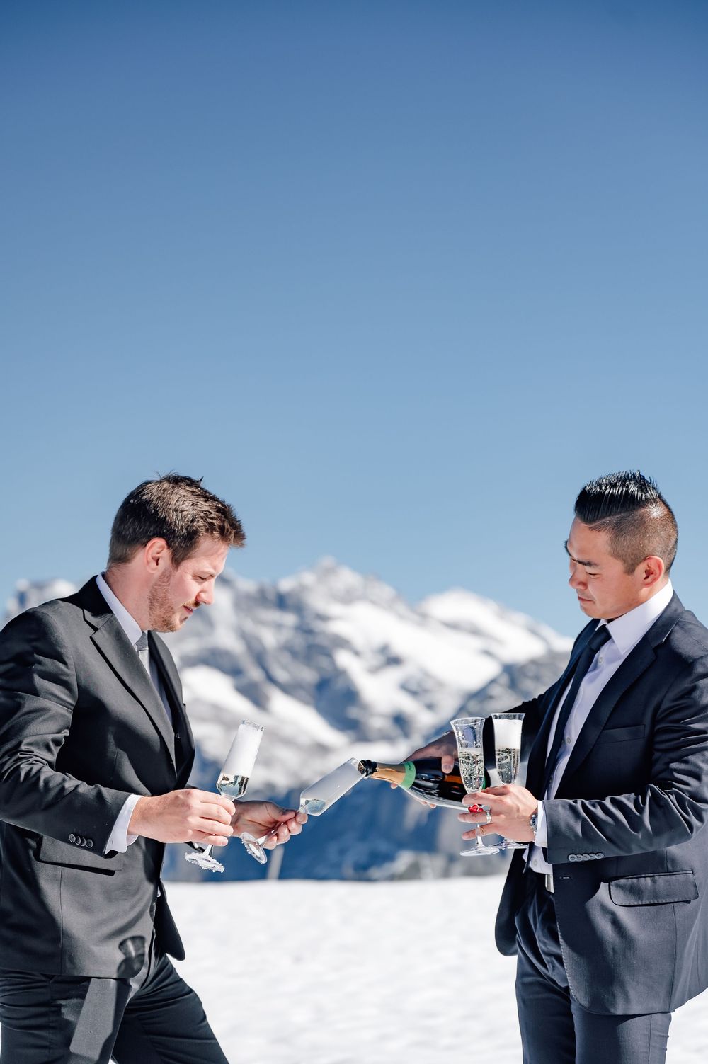 Two businessmen in dark suits toast champagne glasses against a snowy mountain backdrop on a sunny winter day.