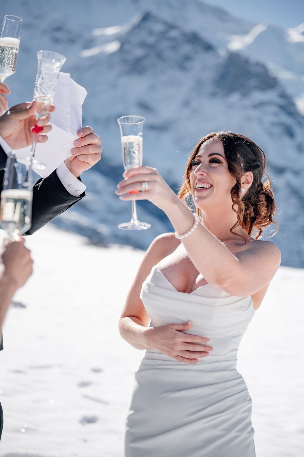 Bride in white dress toasts with champagne against dramatic snow-capped mountain landscape.
