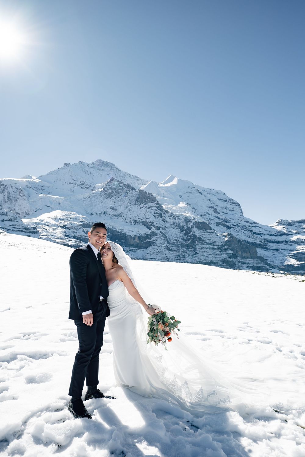 A couple poses for their wedding photos in the snow with majestic snow-capped Swiss Alps mountains in the background.