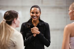 Smiling woman in black jacket stands confidently with clasped hands, engaged in conversation with two others indoors.