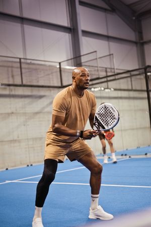 Athletic man in tan outfit playing padel tennis indoors on a blue court, holding a padel racket in ready stance.