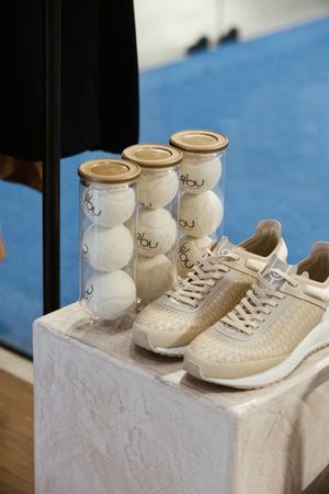 Cream woven sneakers displayed beside three tennis ball canisters labeled rbu on a white pedestal in a retail setting.