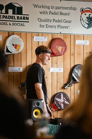 A young man stands smiling beside a speaker at The Padel Barn, surrounded by padel rackets displayed on a wooden wall.