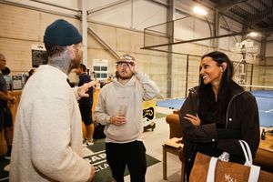 Three people socializing and laughing inside an indoor padel tennis court facility during a casual event.