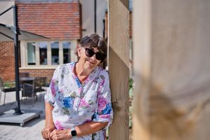 A smiling woman in a floral shirt and sunglasses leans against a wooden post in a sunny outdoor patio setting.