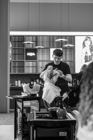 Black and white photo of a hairstylist working on a client in a modern salon with L'Oreal products displayed on shelves.