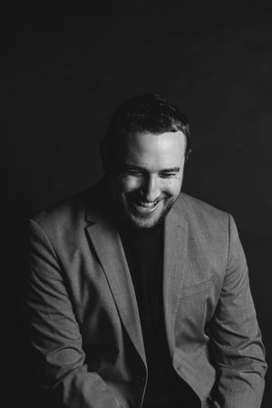 Black and white portrait of a smiling man in a blazer against a dark background.