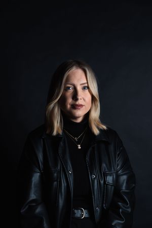 Blonde woman in black leather jacket and turtleneck poses against dark background with gold necklace.