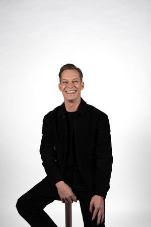 Smiling young man in all-black outfit sitting on a stool against a white background.