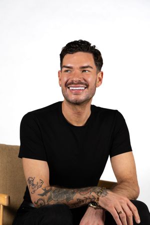 Smiling man with tattoos on his arm wearing a black t-shirt, seated in a chair against a white background.