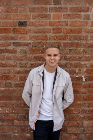 A person wearing a light gray jacket and white t-shirt poses casually against a red brick wall.