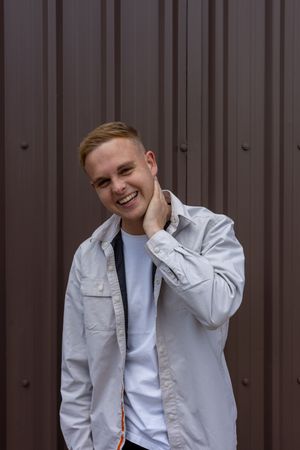 A person in a light gray jacket poses casually against a corrugated metal wall background.