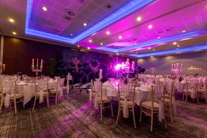 An elegant wedding reception ballroom with purple uplighting, white chairs, and crystal chandeliers creating a romantic ambiance.