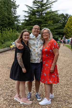 Three friends pose together on a gravel path wearing summer dresses and casual attire in an outdoor garden setting.
