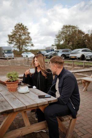 Couple sitting at outdoor wooden picnic table enjoying coffee, surrounded by parked cars and trees on a cloudy day.