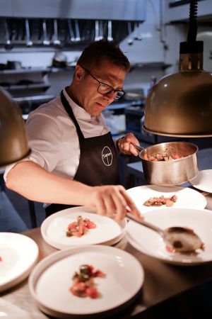 Chef plating gourmet dishes in a professional kitchen, carefully arranging food on white plates under warm lighting.