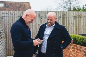 Two men stand by a brick wall while looking at a mobile phone together and sharing a laugh.