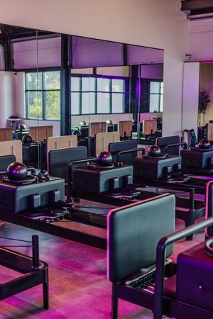 Modern pilates studio with reformer machines arranged in rows under purple lighting and natural daylight.
