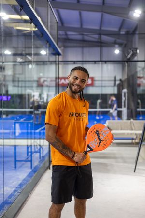 Person in orange t-shirt and black shorts holds a pickleball paddle while smiling on an indoor court.