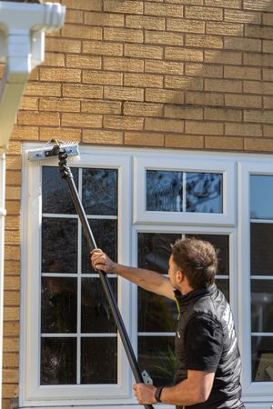 Professional window cleaner using an extension pole to wash exterior windows of a brick house in daylight.