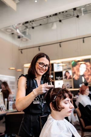 Smiling hairstylist in L'Oreal apron styling a client's short brown hair in a modern salon.