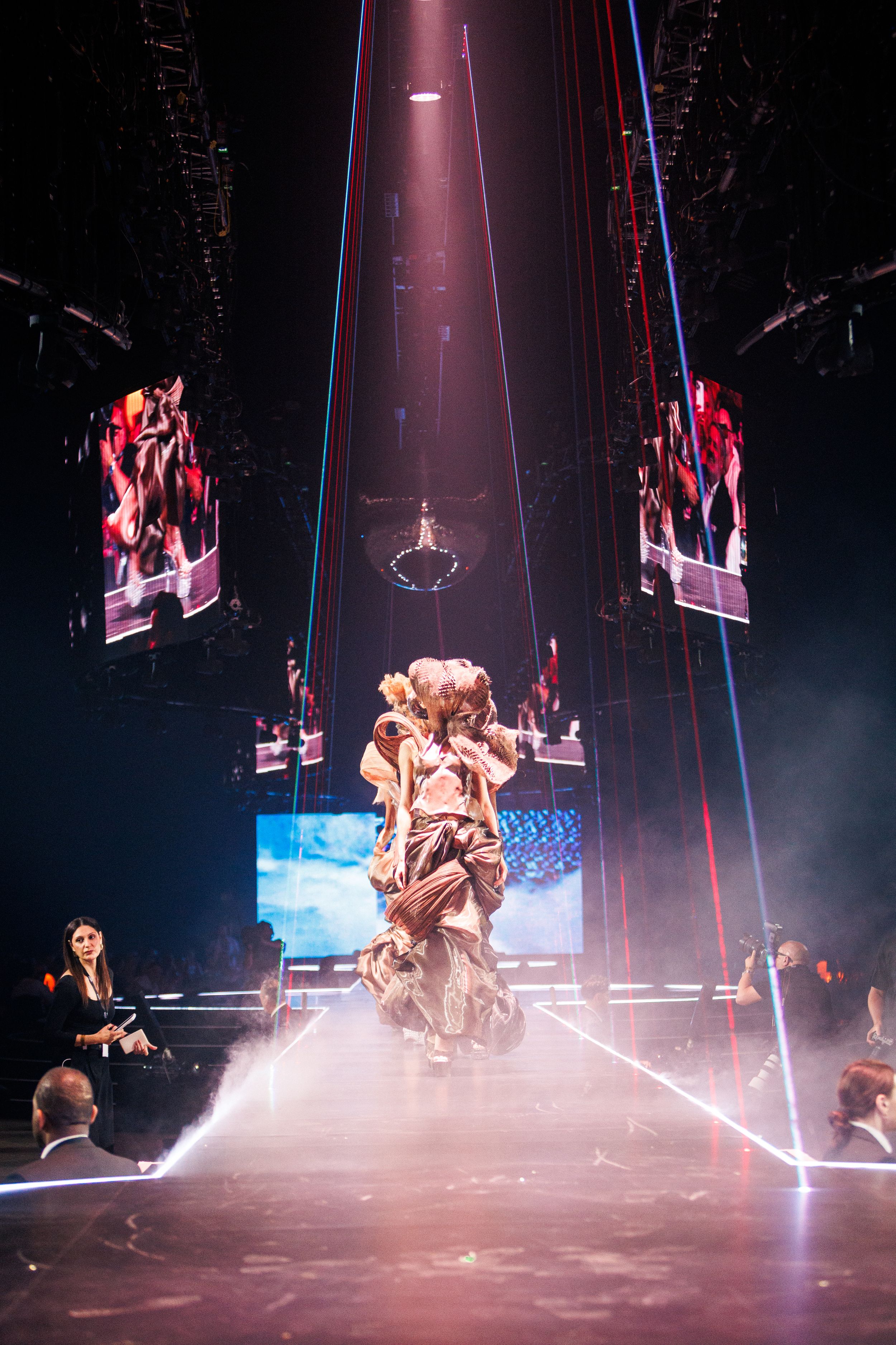 Performer in elaborate costume stands center stage amid laser lights, fog, and large LED screens at a live show.