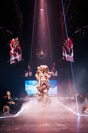 Performer in elaborate costume stands center stage amid laser lights, fog, and large LED screens at a live show.