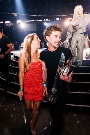 Man holding trophy poses with woman in red dress at awards show venue with stage lights in background.