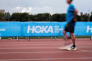 Runner on a red track passing a blue HOKA 'Fly Human Fly' banner at an outdoor athletics event.