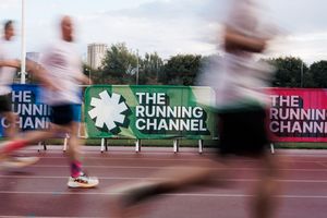 Runners sprint past The Running Channel banner on an athletics track, captured with motion blur effect.