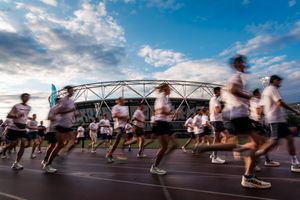 Runners in white shirts race past a large stadium under a dramatic blue sky at dusk during a community running event.