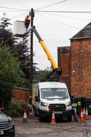 Utility van with yellow crane arm working near brick building with orange traffic cones on residential street.