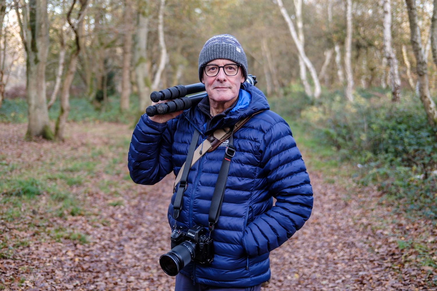 A photographer in a blue puffy jacket and beanie stands on a forest trail with professional camera equipment.